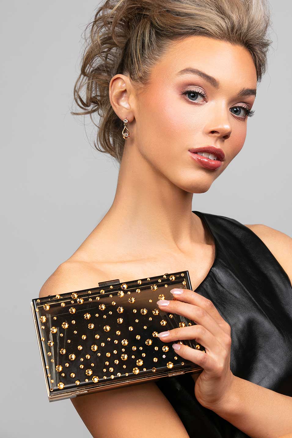 Model wearing the Perry cubic zirconia crystal drop earrings in gold paired with the Parker black lucite clutch that is embellished with gold crystals in a polka dot design
