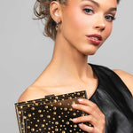 Model wearing the Perry cubic zirconia crystal drop earrings in gold paired with the Parker black lucite clutch that is embellished with gold crystals in a polka dot design