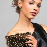 Model wearing the Perry cubic zirconia crystal drop earrings in gold paired with the Parker black lucite clutch that is embellished with gold crystals in a polka dot design