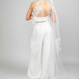 A bride wearing a white chapel veil with floral lace design and Austrian pearl embellishments, posing with her back to the camera.