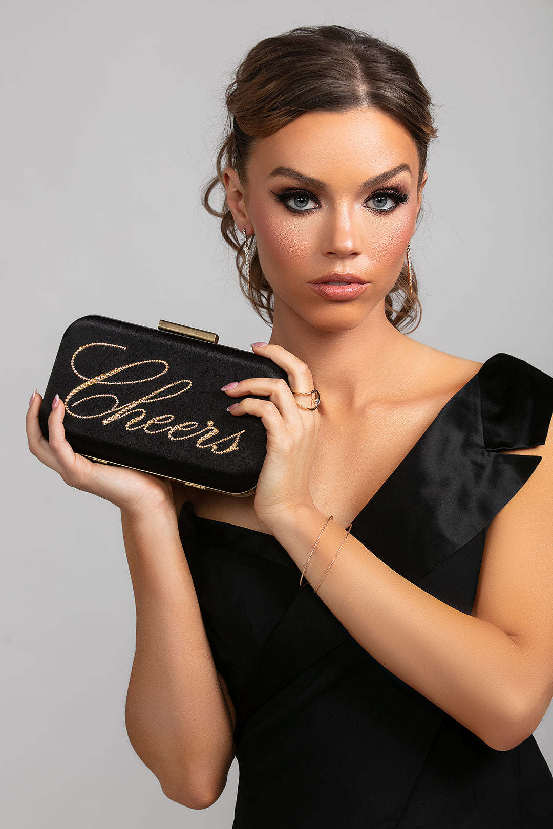 Woman holding a black clutch with 'Cheers' in gold letters against a gray background