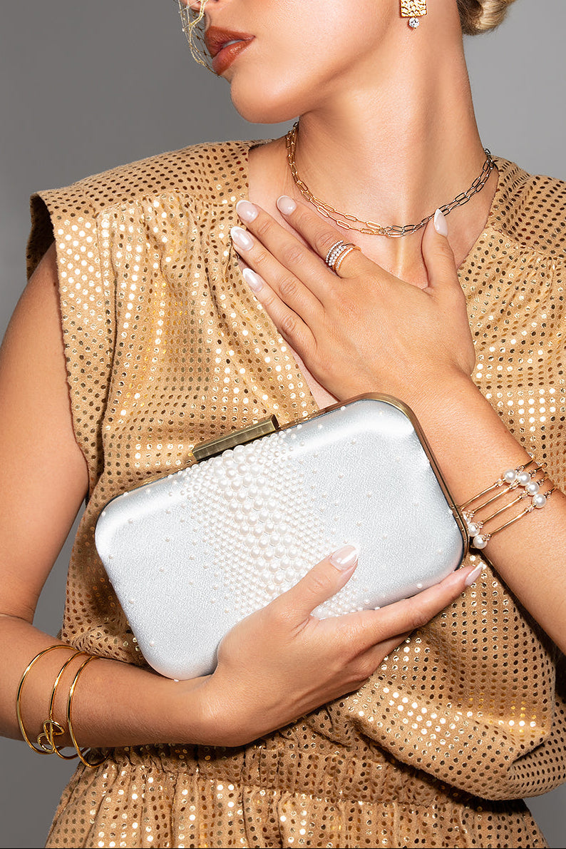 Woman in a gold dress holding an off white pearl clutch against a gray background