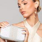 Woman holding a ivory pearl clutch wearing the Taylor gold pearl twist earrings with a sleek bun and gold jumpsuit, against a neutral background
