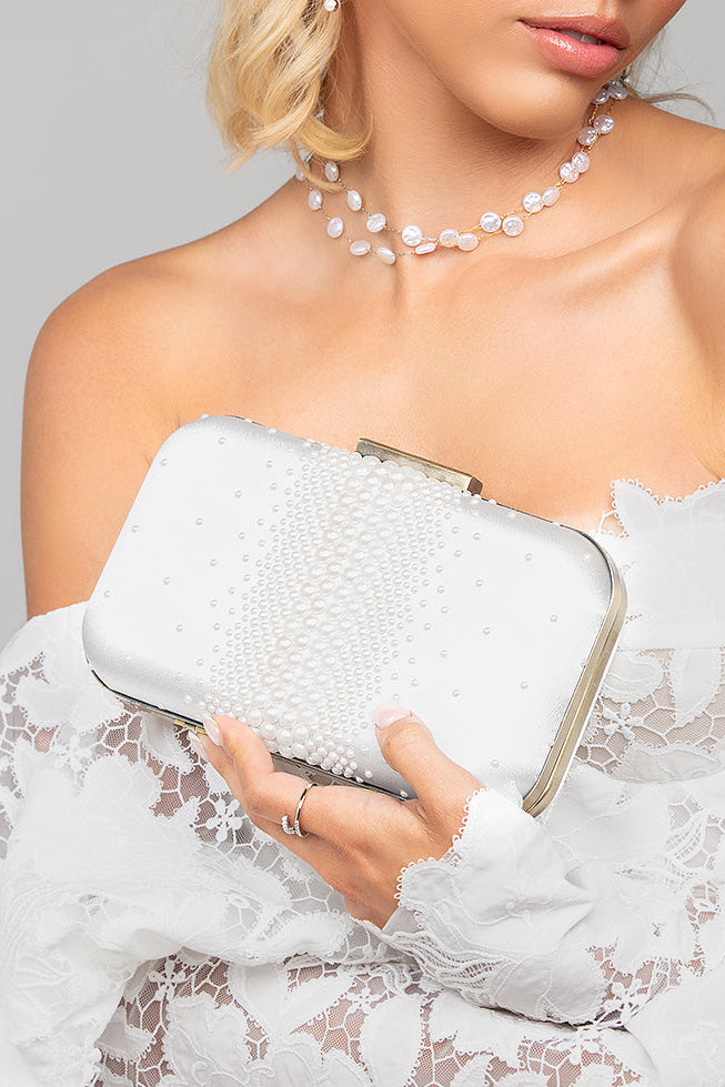 Woman holding a white pearl clutch with a white lacey dress on a plain background