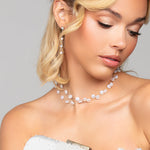 Woman in a white lace dress holding the pearl Tresor clutch by Ariel Taub, paired with the Tori pearl and crystal drop earrings, and Tres Chic pearl choker necklace against a gray background
