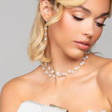 Woman in a white lace dress holding the pearl Tresor clutch by Ariel Taub, paired with the Tori pearl and crystal drop earrings, and Tres Chic pearl choker necklace against a gray background