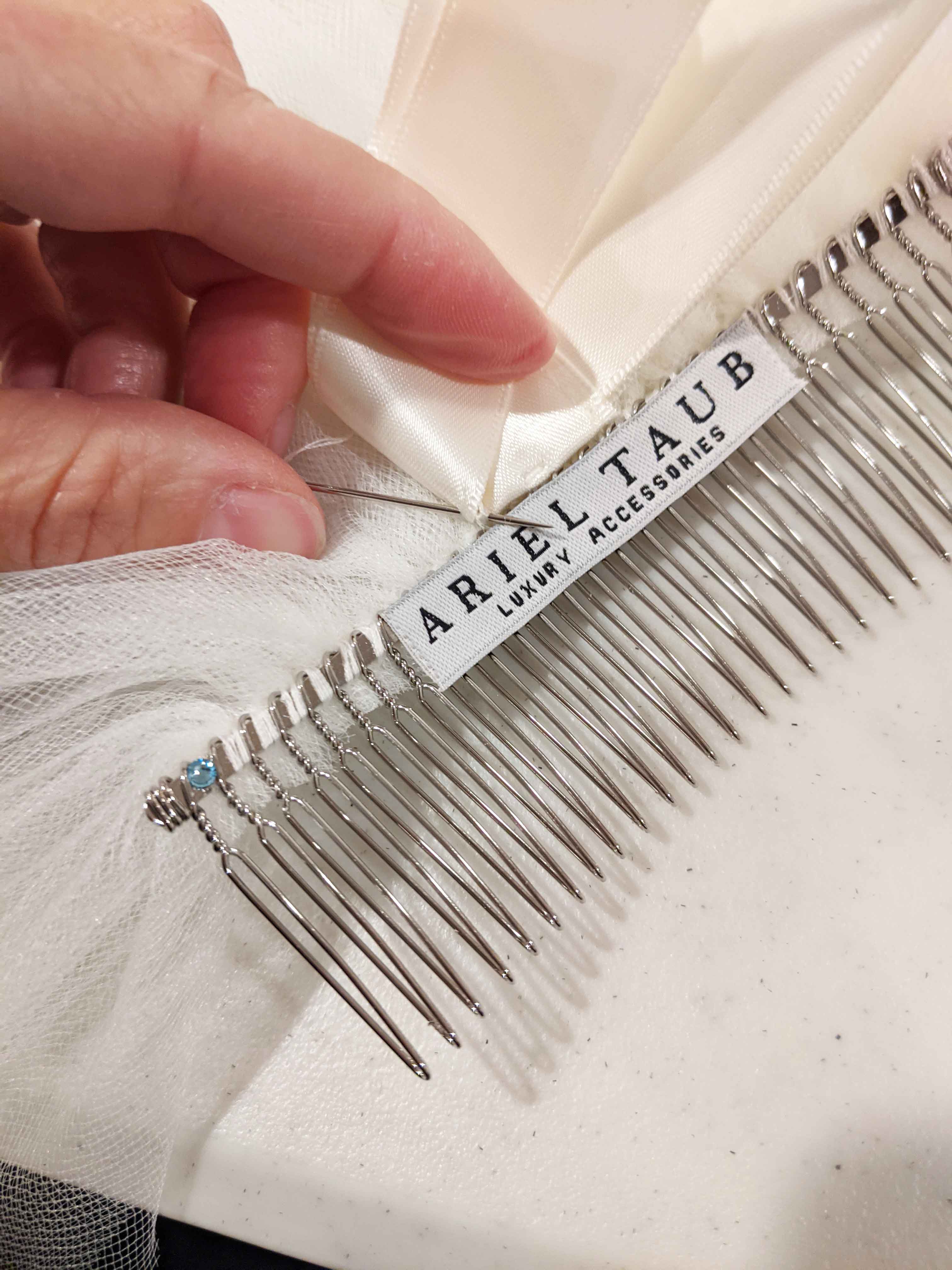 Silver comb being handsewn with 'Ariel Taub' label on a white tulle fabric background and satin ribbon