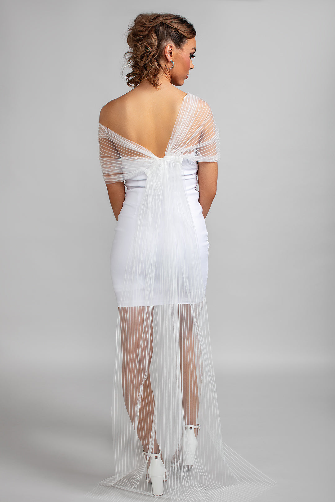 Model wearing the Scarlet off-the-shoulder tulle wrap in off-white, styled with a white strapless mini dress and heels, showing the back view of the sheer striped fabric draping softly across the arms.