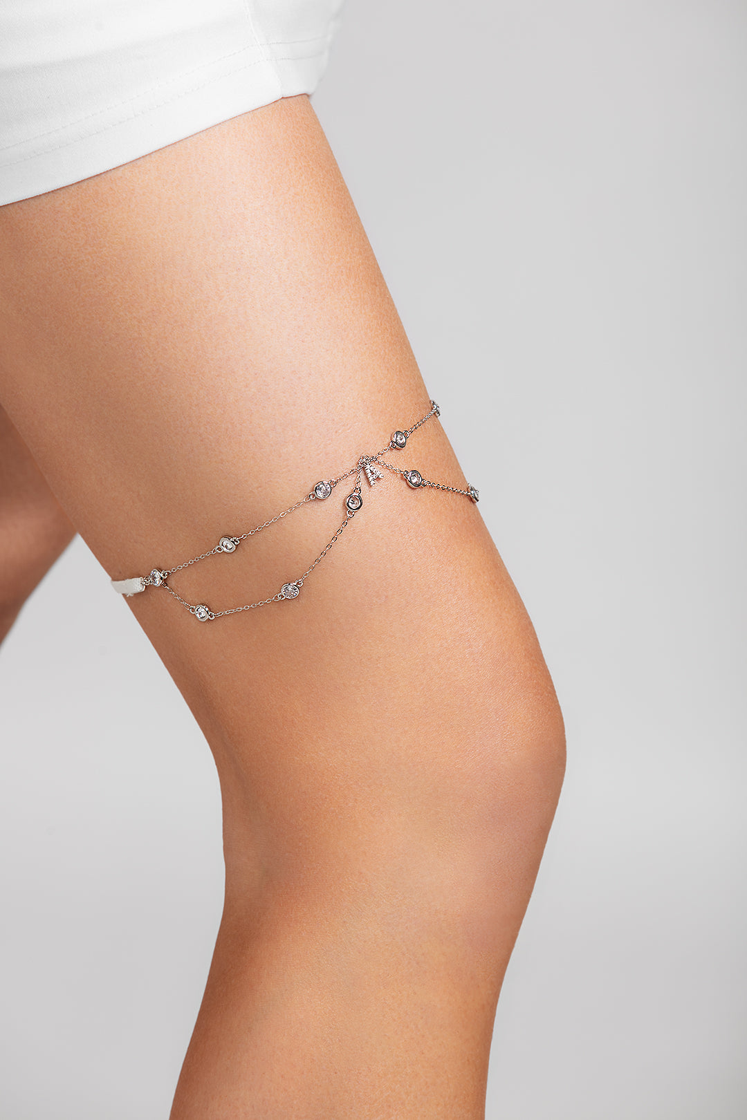 Close-up of a model wearing the Shimmer garter on her thigh, featuring delicate silver chains with crystal accents and a custom crystal initial charm, styled with a white mini dress.