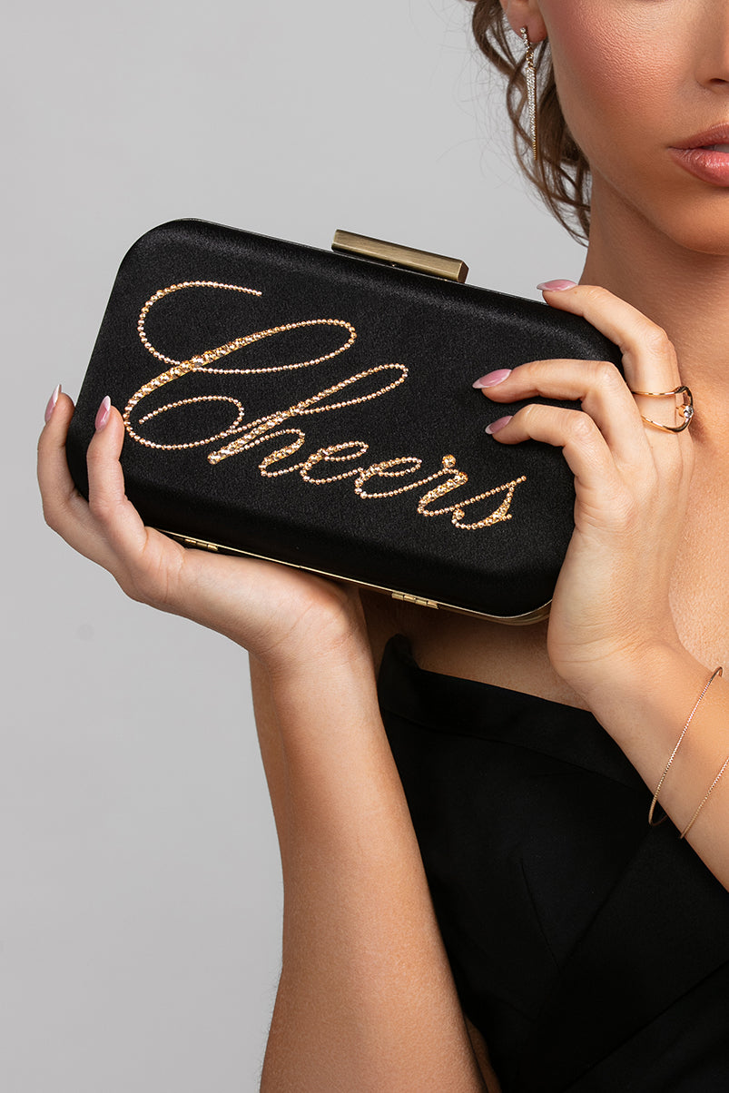 Model holding the Stella clutch with gold "Cheers" script, paired with the Stellar ring and Spencer bracelet in gold.