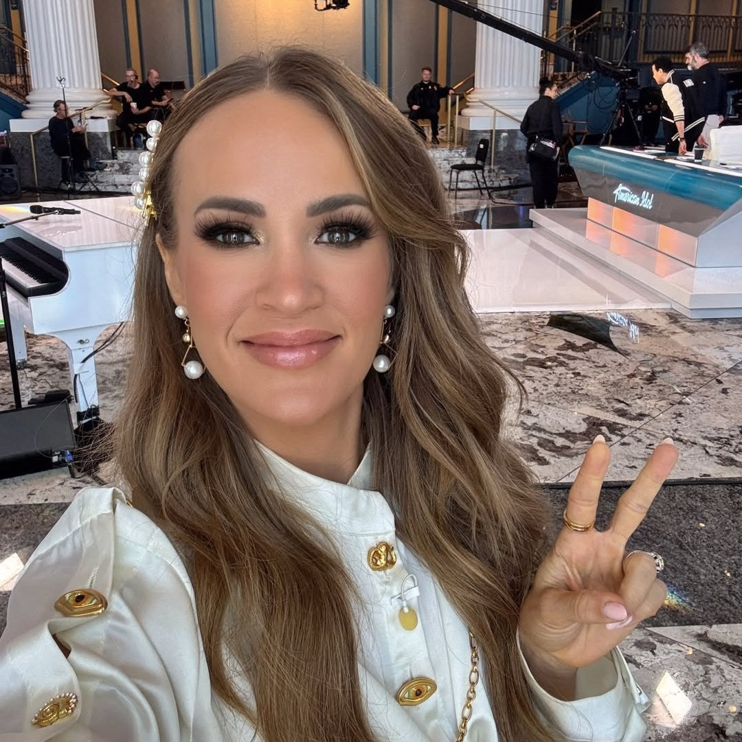 Carrie Underwood in a white outfit with gold buttons making a peace sign in an indoor setting with columns and people in the background. Behind the scenes at American Idol on January 26, 2026 wearing the Ariel Taub Orion earrings and Kimberly hairpiece in gold.