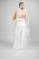 A bride wearing a white chapel veil with floral lace design and Austrian pearl embellishments, posing with her back to the camera.