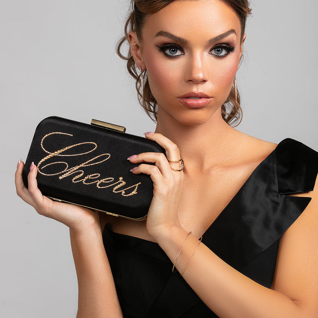 model wearing the Stella spike CZ crystal earrings, Stellar floating CZ crystal ring, and Spencer lightweight bracelet in gold by Ariel Taub. Also holding a black satin clutch with the word Cheers in script made with gold Austrian crystals. 