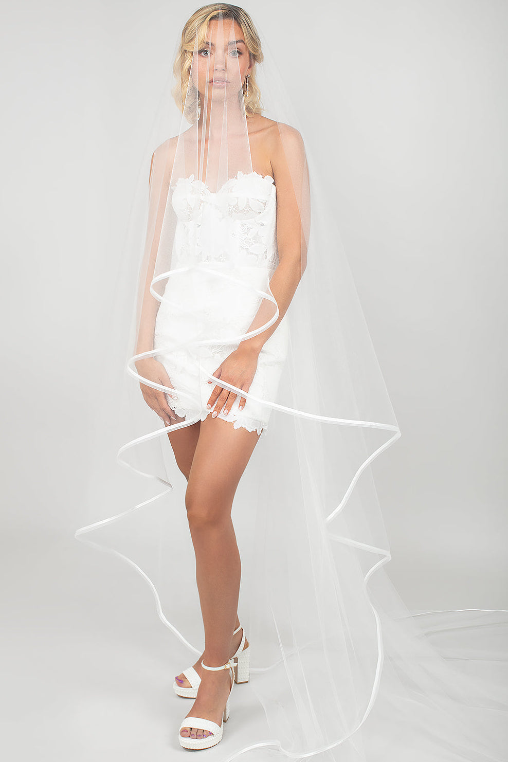 Woman wearing a short white lace dress with a two-tier horsehair and ribbon trimmed wedding veil that is pulled over the face.