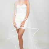 Woman wearing a short white lace dress with a two-tier horsehair and ribbon trimmed wedding veil that is pulled over the face.