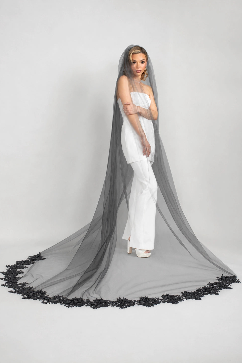Woman in a white strapless pantsuit dress with a long black veil on a gray background