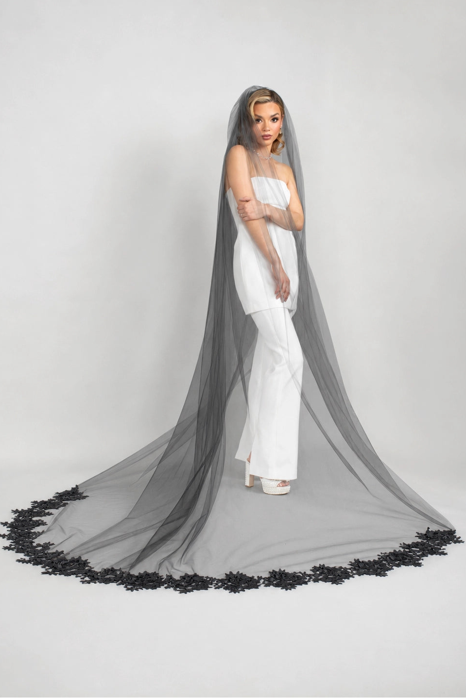Woman in a white strapless pantsuit dress with a long black veil on a gray background