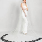 Bride in a white gown with a long veil with black lace rose trim on a gray background