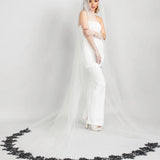 Bride in a white gown with a long veil with black lace rose trim on a gray background
