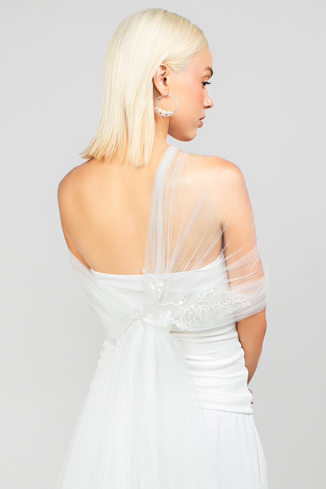 Detail back shot of the Regal tulle wrap with an asymmetrical design and sparkly sequin rose lace applique, adding drama to bridal looks paired with the large Rio Austrian pearl hoop earrings in silver.