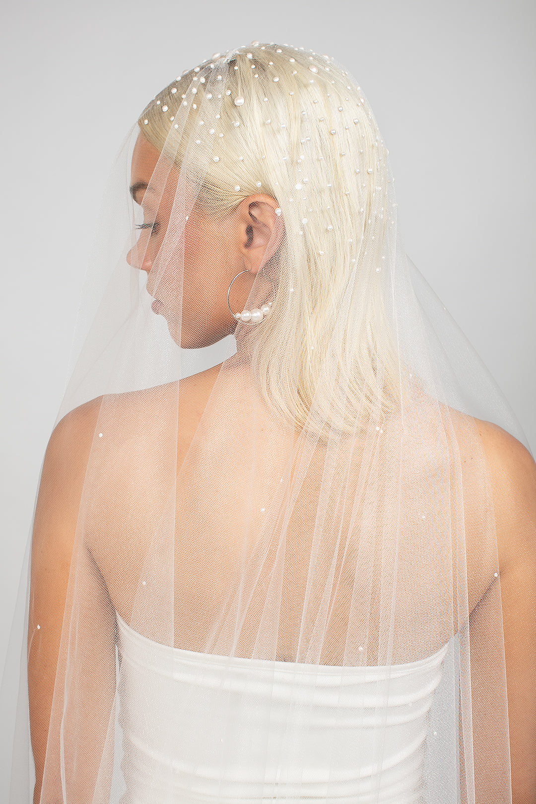 Detail shot of the top of the Royalty bridal veil. Two-tier cathedral-length wedding veil with heavy Austrian pearls clustered at the top, fading into a light scatter toward the bottom, available in multiple shades of white and blush. Paired with the large Austrian hoop Rio earrings in silver.