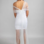 Model wearing the Scarlet off-the-shoulder tulle wrap in off-white, styled with a white strapless mini dress and heels, showing the back view of the sheer striped fabric draping softly across the arms.