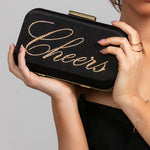 Model holding the Stella clutch with gold "Cheers" script, paired with the Stellar ring and Spencer bracelet in gold.