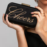 Model holding the Stella clutch with gold "Cheers" script, paired with the Stellar ring and Spencer bracelet in gold.
